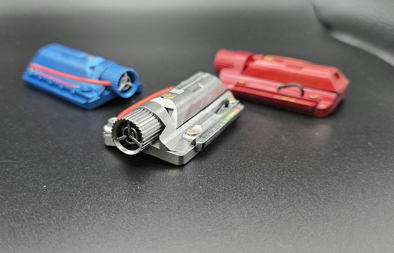 May include: Three miniature engine models in blue, silver, and red. Each rectangular component features a cylindrical intake with a three-spoke fan, exposed wiring, and metallic detailing. The silver model displays small printed text on its side.