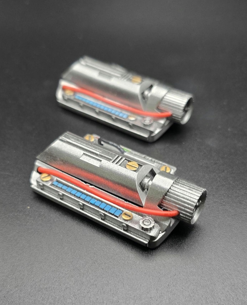 May include: Two rectangular silver mechanical components featuring a metallic finish, a prominent red wire, a row of small blue square accents, and a cylindrical knob on one end. Each piece is secured with small gold-colored screws along the sides.