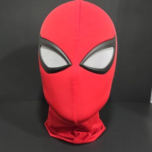 May include: A red Spider-Man mask with black eye frames and white mesh eye coverings. The mask is made of a smooth fabric and covers the entire head and neck. The mask is displayed against a dark background.