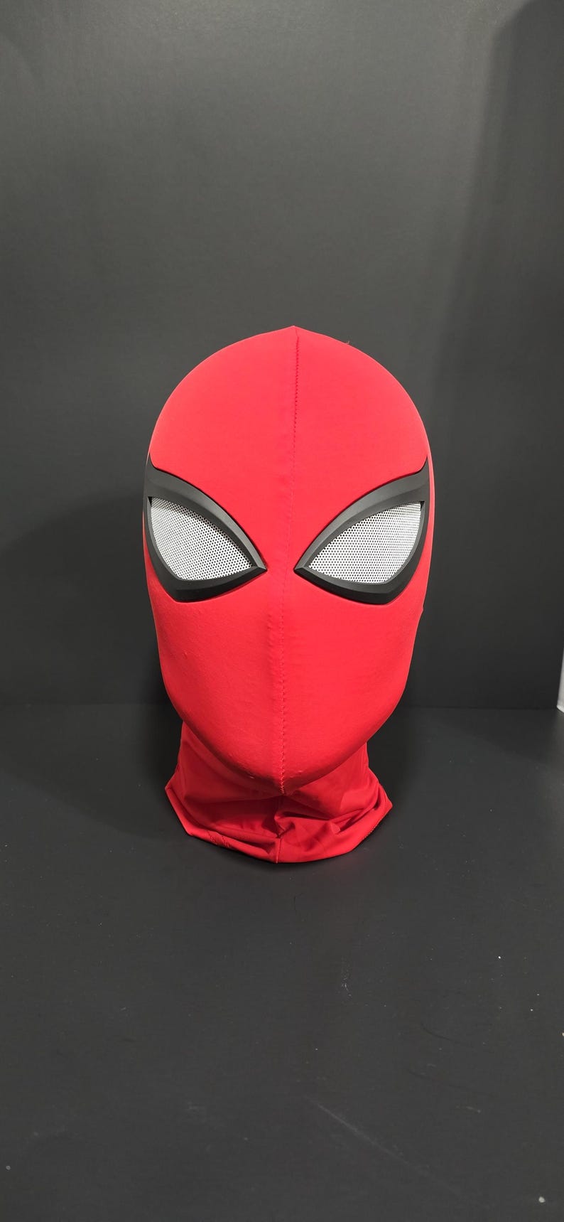 May include: A red Spider-Man mask with black eye frames and white mesh eye coverings. The mask is made of a smooth fabric and covers the entire head and neck. The mask is displayed against a dark background.