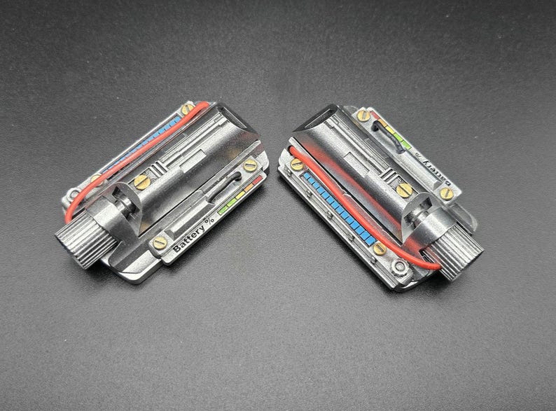May include: Two metallic, rectangular electronic components featuring a silver finish, brass-colored screws, and a red wire. Each unit displays a battery level indicator with the text Battery % and a series of small blue and green square segments.