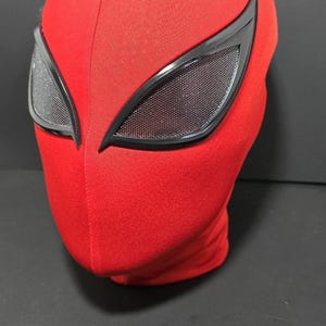May include: A red fabric mask with black-framed eye lenses. The mask covers the entire head and features a textured surface. The eye lenses have a mesh-like appearance.