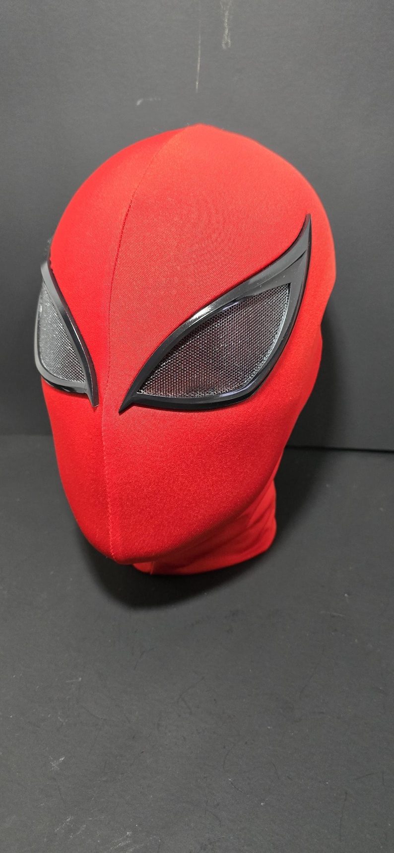 May include: A red fabric mask with black-framed eye lenses. The mask covers the entire head and features a textured surface. The eye lenses have a mesh-like appearance.