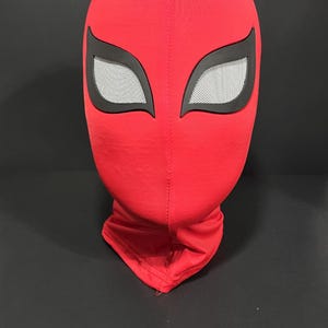 K&ouml;nnte beinhalten: Eine rote Stoffmaske mit schwarzen Augenkonturen und wei&szlig;en Augenabdeckungen aus Mesh. Die Maske ist eine vollst&auml;ndige Kopfbedeckung, wahrscheinlich f&uuml;r Cosplay oder Kost&uuml;me. Die Maske befindet sich auf einer schwarzen Oberfl&auml;che.