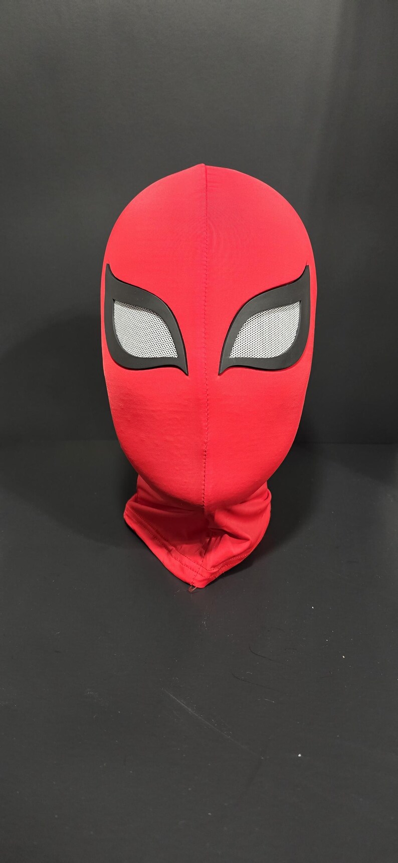 K&ouml;nnte beinhalten: Eine rote Stoffmaske mit schwarzen Augenkonturen und wei&szlig;en Augenabdeckungen aus Mesh. Die Maske ist eine vollst&auml;ndige Kopfbedeckung, wahrscheinlich f&uuml;r Cosplay oder Kost&uuml;me. Die Maske befindet sich auf einer schwarzen Oberfl&auml;che.