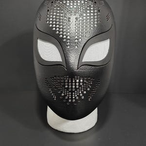K&ouml;nnte beinhalten: Schwarze Maske mit strukturierter Oberfl&auml;che und Mesh-Augenabdeckungen. Die Maske hat eine Reihe kleiner, gleichm&auml;&szlig;ig verteilter L&ouml;cher oben und unten. Die Maske ruht auf einer wei&szlig;en Schaumbasis.