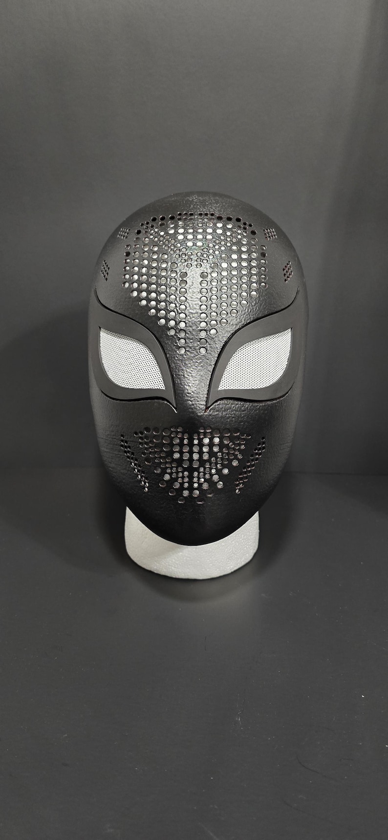 K&ouml;nnte beinhalten: Schwarze Maske mit strukturierter Oberfl&auml;che und Mesh-Augenabdeckungen. Die Maske hat eine Reihe kleiner, gleichm&auml;&szlig;ig verteilter L&ouml;cher oben und unten. Die Maske ruht auf einer wei&szlig;en Schaumbasis.