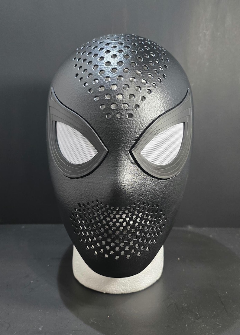 May include: Black Spider-Man mask with a textured surface and eye-shaped openings. The mask has a perforated design around the mouth and top of the head, and is likely a costume accessory.