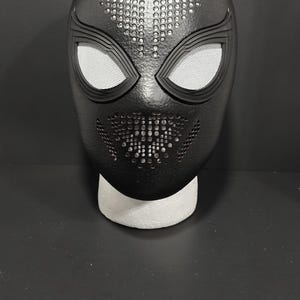 May include: Black superhero mask with white mesh eye coverings and a detailed, textured design. The mask has a perforated top and a stylized mouth area, resting on a white foam base. A striking costume accessory.