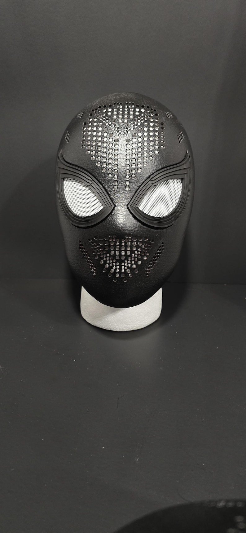 May include: Black superhero mask with white mesh eye coverings and a detailed, textured design. The mask has a perforated top and a stylized mouth area, resting on a white foam base. A striking costume accessory.