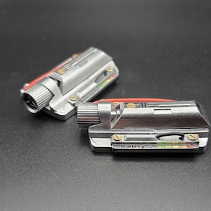 May include: Two metallic silver rectangular components featuring a cylindrical knob on one end, gold-colored screws, and a small display panel labeled Battery % with a multi-colored indicator bar. A thin black wire and a red accent piece are visible.