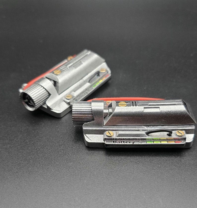 May include: Two metallic silver rectangular components featuring a cylindrical knob on one end, gold-colored screws, and a small display panel labeled Battery % with a multi-colored indicator bar. A thin black wire and a red accent piece are visible.