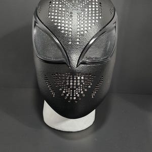 May include: Black full-face mask with a textured surface and a perforated design. The mask features eye openings with mesh and a pattern of small holes across the top and lower sections. The mask is displayed on a white stand.