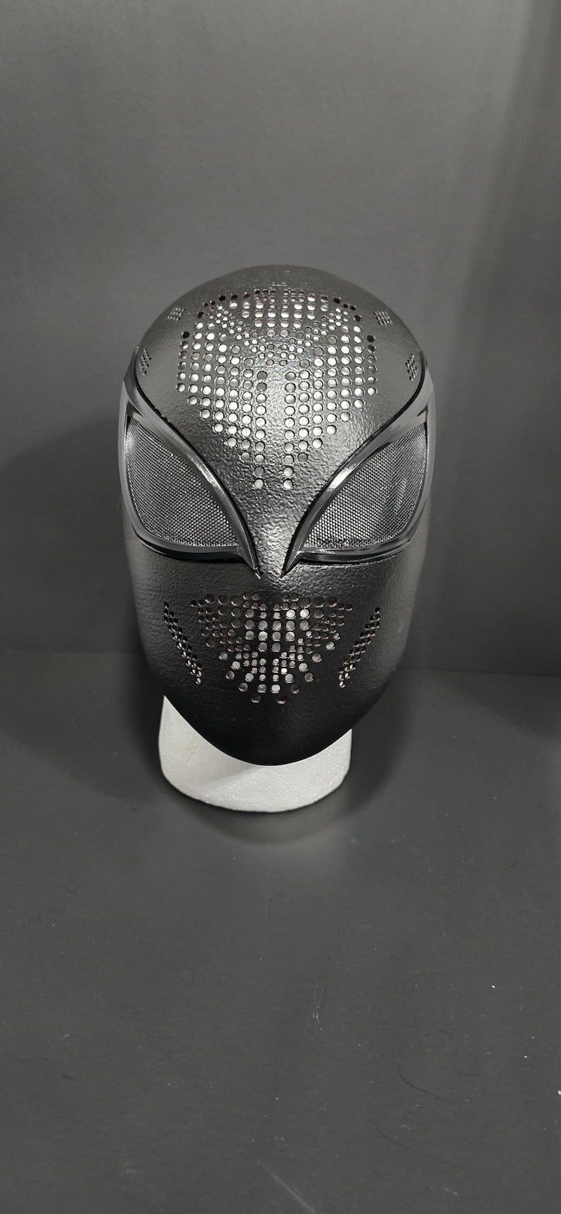 May include: Black full-face mask with a textured surface and a perforated design. The mask features eye openings with mesh and a pattern of small holes across the top and lower sections. The mask is displayed on a white stand.
