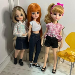 May include: Three fashion dolls with different hairstyles and outfits. One doll wears a white sequined hoodie and denim skirt, another wears a white crop top and jeans, and the third wears a pink plaid top and black shorts.