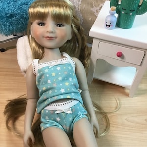 May include: A doll wearing a blue and white star patterned tank top and matching underwear. The doll has long blonde hair and is lying on a wooden floor.