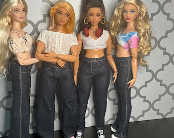 May include: Four fashion dolls wearing denim jeans and crop tops. The dolls have different hairstyles and skin tones. One doll has headphones around their neck. The dolls are standing in front of a patterned background.