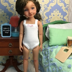 May include: A doll wearing a white sleeveless bodysuit with a pink bow on the neckline. The doll has long brown hair and blue eyes. The doll is standing in front of a green bed with a white and green patterned blanket. The doll is holding a sign that says "Welcome to OG's garden Party!"