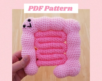 May include: A pink crocheted plush toy shaped like a human intestine. The toy has a smiling face with two black eyes and a mouth. The text "PDF Pattern" is at the top of the image. The text "CraftWithCrohns" is at the bottom of the image.