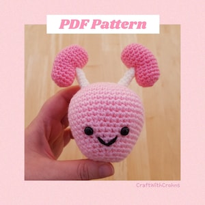 Op de afbeelding: Een roze gehaakt amigurumi-speelgoed met witte antennes en zwarte ogen. Het speelgoed heeft een glimlachende mond en wordt in een hand gehouden. De afbeelding bevat de tekst "PDF Pattern" en "CraftWithCrohns".