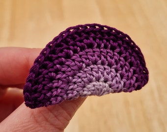 May include: A purple and white crocheted flower with a gradient of color. The flower is made with a single crochet stitch and has a scalloped edge.