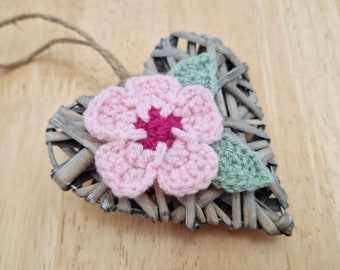 May include: A woven heart-shaped ornament with a pink crocheted flower and green crocheted leaves.