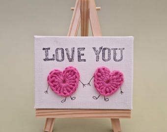 May include: A small white canvas with the words "I LOVE YOU" printed on it. Two pink crocheted hearts with black drawn legs and arms are holding hands. The canvas is on a small wooden easel.