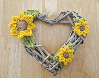 May include: A heart-shaped wicker wreath decorated with three crocheted sunflowers. The sunflowers are yellow with brown centers and green leaves.