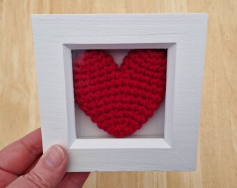 May include: A white frame with a red crocheted heart inside. The heart is framed with a clear glass panel.