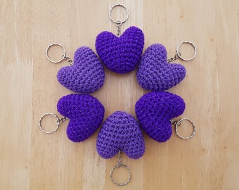 May include: Six purple crocheted heart-shaped keychains with silver key rings. The keychains are arranged in a circle on a wooden surface.