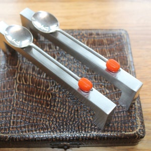 May include: A pair of silver spoons with rectangular handles and orange accents, resting on a brown, textured box. The spoons have small, round bowls and are designed with a modern aesthetic.