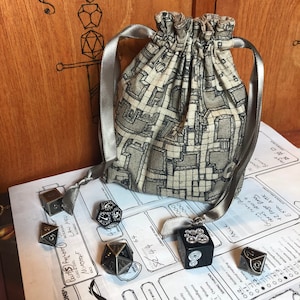 May include: A white and grey fabric drawstring bag with a dungeon map design. The bag is filled with silver metal dice. The dice are scattered on a white surface with black text.