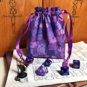 Peut inclure: Un sac à dés en tissu violet avec un motif géométrique. Le sac est attaché avec un ruban violet et est posé sur une table avec des dés éparpillés autour.
