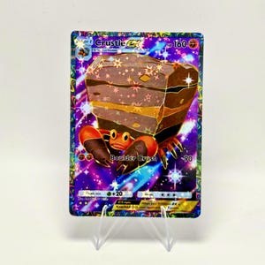 May include: A Crutle EX Pokemon trading card with a colorful design. The card features a large, brown rock-like creature with orange claws and a purple and starry background. The card is displayed on a clear stand.