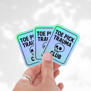 May include: Three holographic stickers with a teal and purple gradient. Each sticker has a black border and the text "TOE PICK TRAUMA CLUB" with a skull graphic. The stickers are held in a hand.