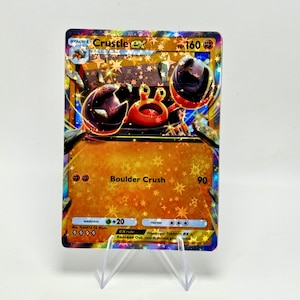 May include: A holographic Crustle EX Pokémon card. The card features a red and brown crab-like creature with the text "Crustle EX" and "Boulder Crush". The card is displayed on a clear plastic stand.