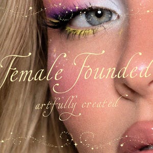 Puede incluir: Un primer plano de un rostro con cabello rubio, ojos azules y maquillaje vibrante con sombras de ojos moradas y amarillas. El texto "Female Founded" y "artfully created" está superpuesto en una fuente cursiva dorada con destellos decorativos.