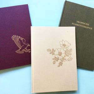 May include: Three hardcover books with gold foil designs. One is deep purple with an eagle, another is cream with flowers, and the third is olive green with the text "TROPISCHE SCHMETTERLINGE".