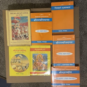 Może przedstawiać: Kolekcja książek religijnych z pomarańczowymi i żółtymi okładkami. Tytuły obejmują "Bhagavad-Gita As It Is" i "Shrimad Bhagavadgita". Książki zawierają tekst w wielu językach i ilustracje.