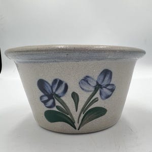 Puede incluir: Una maceta de cerámica gris con un diseño floral azul. La maceta tiene una forma ligeramente acampanada y un borde azul claro. Dos flores azules con tallos y hojas verdes están pintadas en la parte delantera.