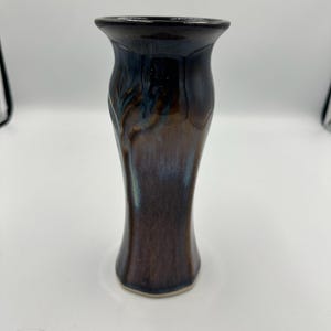 Może przedstawiać: Wysoki, ceramiczny wazon z rozszerzoną górą i wąską podstawą. Wazon ma błyszczące wykończenie z mieszanką ciemnobrązowych, niebieskich i brązowych odcieni. Projekt ma subtelny, organiczny wzór.