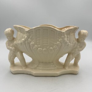 May include: Cream-colored ceramic planter with a scalloped rim and a decorative shell-like design. Two cherubic figures support the planter on either side. The planter has a rectangular base.