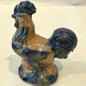 May include: A ceramic cockerel figurine featuring a glossy finish with a two-tone colour scheme. The body is primarily a light tan or beige colour, while the comb, beak, neck feathers, wings, and tail feathers are accented in a deep, dark blue glaze.