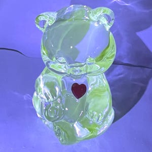 May include: A translucent green glass bear figurine with a red heart on its chest. The bear has visible ears, arms, and legs. The glass has a slight yellow tint, and the figurine is sitting on a blue surface.
