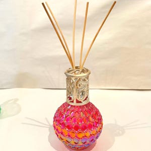 May include: A decorative glass diffuser with a round, textured, pink and orange body. The diffuser has a silver-colored, openwork top and contains several wooden reeds. The diffuser is designed for home fragrance.