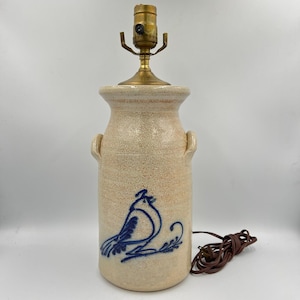 Puede incluir: Una lámpara de mesa de cerámica con un acabado beige moteado y un portalámparas de latón. La base de la lámpara presenta un diseño de pájaro azul. La lámpara tiene dos asas pequeñas y un cable eléctrico marrón.