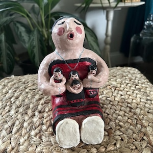 May include: A handmade ceramic figurine of a woman holding three children. The figurine is painted with red, black, and white details. The woman has rosy cheeks and is wearing a necklace. The figurine is sitting on a woven surface.