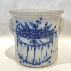 Puede incluir: Un tarro de gres con una base beige claro y un diseño floral azul. El diseño presenta una cesta de bayas y hojas. El tarro tiene dos asas y un borde ancho. El diseño general es rústico y vintage.