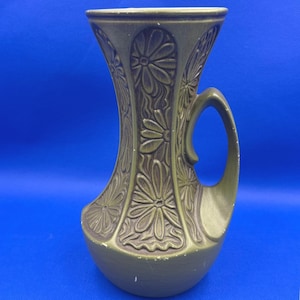 May include: An olive green ceramic pitcher featuring a flared rim and a single handle. The exterior surface is decorated with a repeating embossed floral pattern consisting of stylized daisy-like flowers arranged in vertical panels around the body.