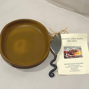 Può includere: Un piatto rotondo in ceramica marrone con manico, abbinato a una spatola per formaggio in metallo con manico nero ondulato legato con spago. Accanto si trova una scheda ricetta intitolata VERMONT APPLE MAPLE BRIE MELT di BENNINGTON POTTERS.